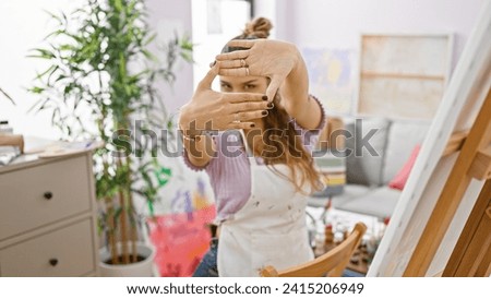 Cheery young hispanic woman artist snapping whimsical photo gesture with hands, radiating beauty and bubbling creativity in vibrant art studio