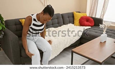 Worried african american woman nursing painful leg injury, sitting on sofa at home