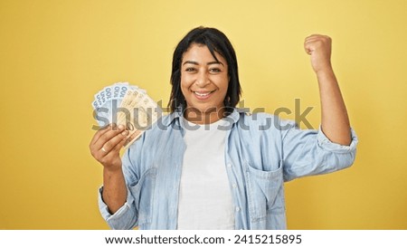 A joyful woman holding brazilian currency flexes her arm against a yellow background, symbolizing financial success. Royalty-Free Stock Photo #2415215895
