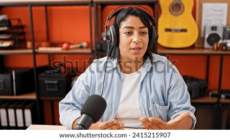 Hispanic woman podcasting in a music studio with headphones, microphone, and guitar background.