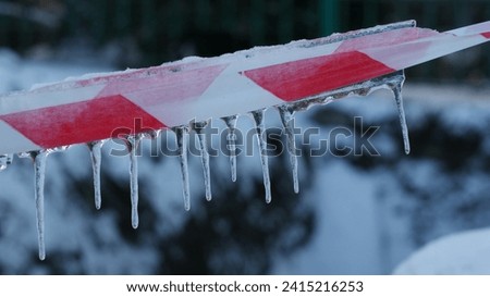 Caution tape, covered in snow and icicles. The tape is stretched horizontally, with icicles hanging from its bottom edge against a blurred snowy landscape Royalty-Free Stock Photo #2415216253