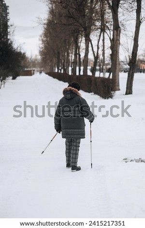 An adult, elderly woman with aluminum hiking sticks walks along the road in winter on white snow on the street outdoors. Photography, healthy lifestyle concept, outdoor sports.