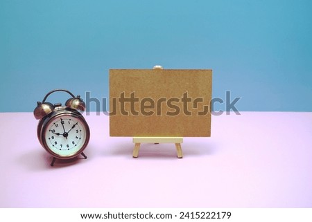 Empty card mockup space for text message with wooden easel and vintage alarm clock on pink background, business and education background