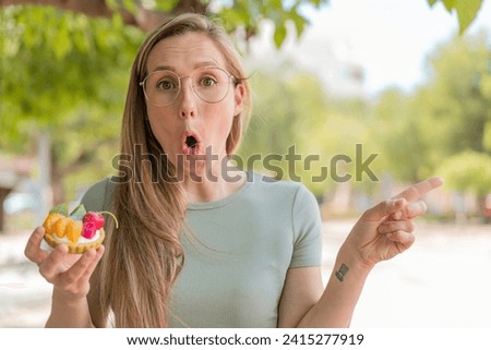 Young blonde woman holding a tartlet at outdoors surprised and pointing side