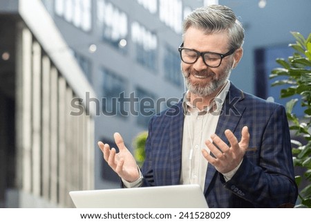 Mature professional man in business attire engaged in a video call using a laptop outdoors with an office backdrop. Royalty-Free Stock Photo #2415280209