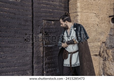 A medieval swordsman in traditional attire stands by an old castle gate, holding his sword. Royalty-Free Stock Photo #2415285473