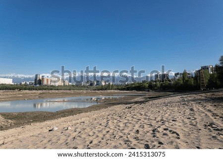Storage reservoir Lake Sayran, Almaty, Kazakhstan. Empty City sand beach with drained pond. Residential apartment buildings and high snow-capped mountains in background. Recreation place for citizens Royalty-Free Stock Photo #2415313075