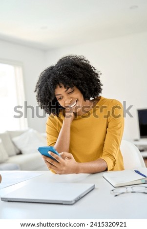 Happy smiling young African woman customer sitting at home table looking at cellphone while holding mobile cell phone in hands texting using smartphone scrolling media, shopping online, vertical.