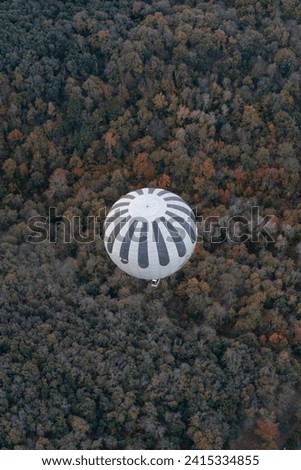 View from above of a hot air balloons flying over pinewood forest at La Garrotxa Volcanic Natural Park. Hot air balloon with black and white stripes with autumnal landscape in the background.