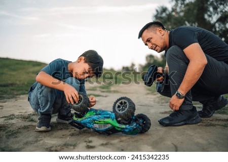 Picture of dad and his son crouching next to overturned toy car. They are looking at the car since it is broken. Father is holding a remote controller, boy is checking the wheel. Blurred foreground.