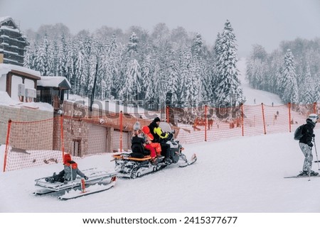 Dad and children ride a snowmobile with a sled attached on a snowy mountain along a red fence