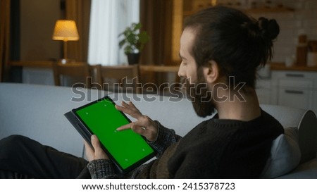 Bearded man scrolling and tapping center on tablet with green screen mock up display. Male lyin on sofa, relaxing at home. Close-up over the shoulder shot