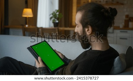 Bearded hair bun man scrolling and tapping center on tablet with green screen mock up display. Male lyin on sofa, relaxing at home. Close-up over the shoulder shot
