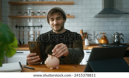 Smiling man with beanie sitting in modern kitchen at home while holding smartphone in his hand looking at the camera and puts couple of coin into the piggy bank