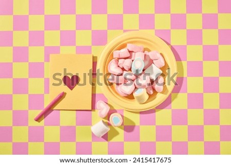 Against the pink and yellow checkered background, a yellow plate filled with sweet marshmallow decorated with a yellow paper card. Top view, creative scene for advertising, valentine’s day
