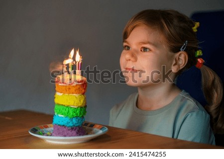 Happy little preschool girl celebrating birthday. Closeup of child with homemade rainbow cake, indoor. Happy healthy toddler blowing six candles on cake. Selective focus on cake