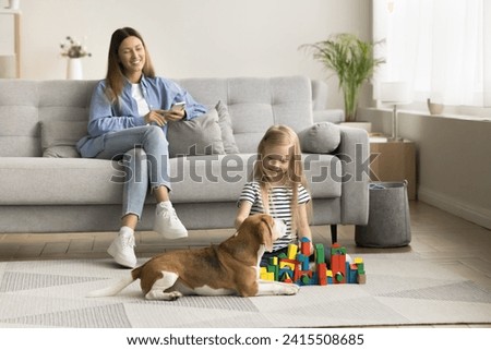 Happy little kid playing with cute beagle dog and stacking building blocks on warm floor. Positive mom using cellphone, looking at little daughter from comfortable couch on family weekend Royalty-Free Stock Photo #2415508685