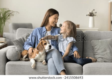 Happy beautiful mother embracing kid and touching dog on home sofa, talking to little daughter girl, smiling, laughing, relaxing in cozy home interior, spending family leisure with kid and pet Royalty-Free Stock Photo #2415508693