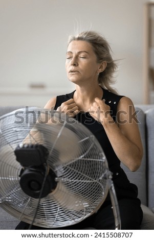 Vertical shot mature woman suffers from unbearable hot temperature inside living room without air-conditioner, cooling, use electric ventilator at home on hot summer day, feel unwell, looking stressed Royalty-Free Stock Photo #2415508707
