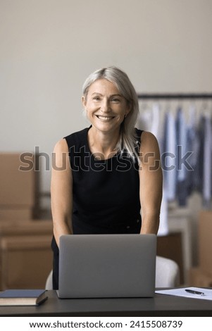 Portrait of middle-aged small business owner posing in warehouse. Female designer standing at desk with laptop at personal atelier workplace. Entrepreneurship, distribution, dropshipping, workflow Royalty-Free Stock Photo #2415508739