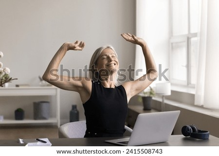 Happy middle aged business woman sitting at desk with eyes closed, rising hands, stretch back after workday finishing, satisfied with good job done, plan weekend, feel successful completion of project Royalty-Free Stock Photo #2415508743
