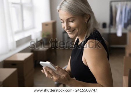 Close up shot of smiling mature 45s woman using smartphone working in warehouse, make call to delivery services, reliable transporting company on relocation day, stacked cardboard boxes on background Royalty-Free Stock Photo #2415508747