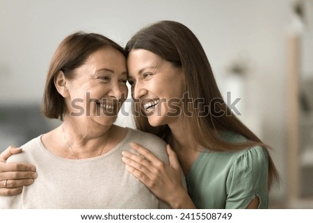 Happy young adult daughter woman hugging elder mature mom from behind, touching mothers shoulders, talking, laughing with head touches, enjoying family bonding, parent and child relationship Royalty-Free Stock Photo #2415508749