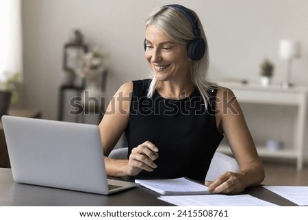 Happy pretty mature woman in headphones sit at desk, take notes, watch webinar on laptop, enjoy web course or online training, study remotely from home office, talking on video call to business client Royalty-Free Stock Photo #2415508761