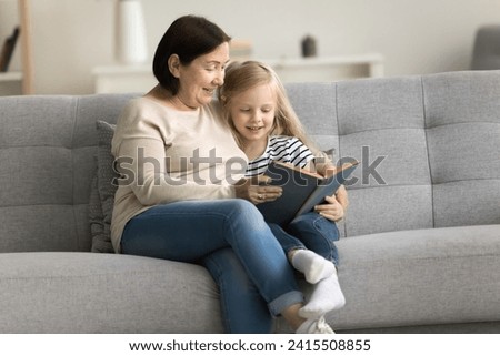 Positive caring grandma reading book to granddaughter kid on comfortable couch. Grandmother telling fairytale to little girl at home, teaching child to read, enjoying educational activity