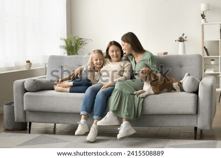 Wide family home portrait of three female generations with dog. Happy grandma, mother and little kid girl stroking cute beagle, sitting on sofa close together, talking, laughing Royalty-Free Stock Photo #2415508889