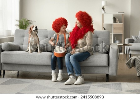 Cheerful little kid girl and grandma in red wigs sitting on couch at trick dog, playing toy drum, singing to music, smiling, laughing, having fun with pet at home masquerade party Royalty-Free Stock Photo #2415508893
