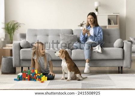 Happy mom taking picture of pet and child, sitting on comfortable sofa, watching little kid and dog playing with building cubes on warm floor, using mobile phone, shooting, recording