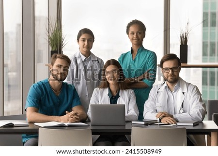 Positive diverse team of attractive young practitioners, surgeons, nurses posing together in hospital office, sitting and standing at table with laptop, looking at camera for portrait