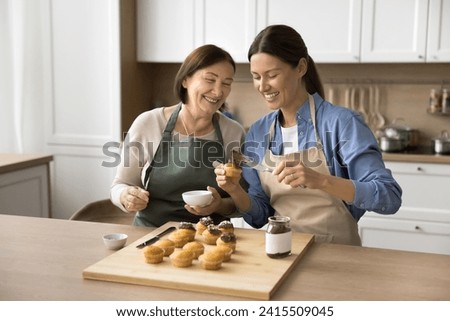 Cheerful senior mom and adult daughter woman in aprons enjoying baking activity in home kitchen, decorating muffins, cupcakes with chocolate creamy cap, preparing homemade dessert for party Royalty-Free Stock Photo #2415509045