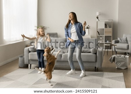 Excited mom and happy preschool daughter kid dancing with playful pet, hopping in living room, having fun, laughing, enjoying active leisure, music at home, training adorable beagle dog. Full length Royalty-Free Stock Photo #2415509121