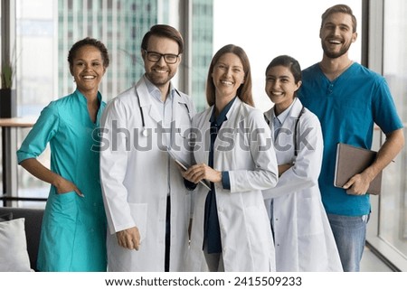 Multiethnic team of happy cheerful young practitioners, surgeons, nurses standing together in hospital office hall, looking at camera, laughing, posing for group portrait