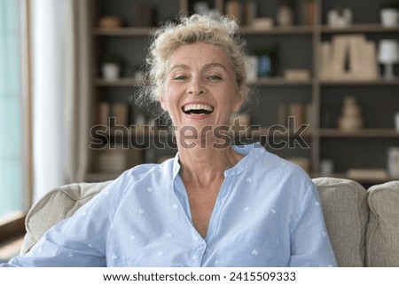 Head shot portrait of happy middle-aged woman, sitting on cozy couch in living room laughing, staring at camera, enjoy carefree retired life alone at home. Midlife, optimistic mood, videocall event Royalty-Free Stock Photo #2415509333