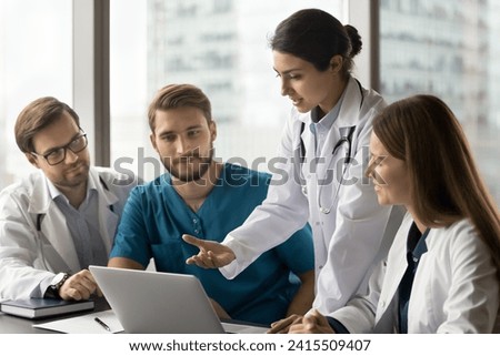 Young Indian doctor woman showing online service for practitioners, speaking at laptop. Multiethnic medical colleagues discussing healthcare application, looking at computer screen at workplace Royalty-Free Stock Photo #2415509407