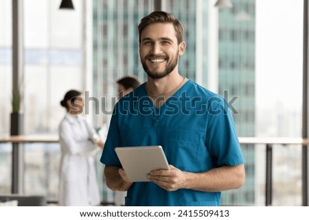 Cheerful handsome surgeon doctor man in blue uniform holding digital tablet computer, looking at camera, smiling, posing for portrait in clinic hall, promoting modern technology in medical job Royalty-Free Stock Photo #2415509413