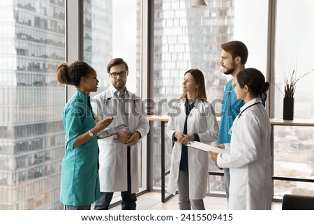 City hospital diverse staff of young practitioners in uniforms standing in office hall, talking, meeting for teamwork at large window with urban view, discussing profession, consulting colleagues Royalty-Free Stock Photo #2415509415