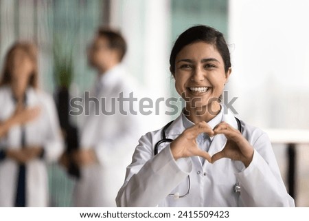 Cheerful young Indian doctor woman making hand heart, looking at camera with happy toothy smile, showing romantic gesture of love, care, health protection, cardiology healthcare Royalty-Free Stock Photo #2415509423