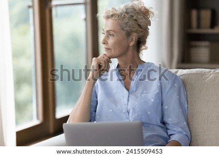 Serious 55s woman sit on sofa with laptop, deep in thoughts looks into distance, thinking over problem, solve difficult task, faced up with modern app lack of understanding. Older gen and modern tech Royalty-Free Stock Photo #2415509453