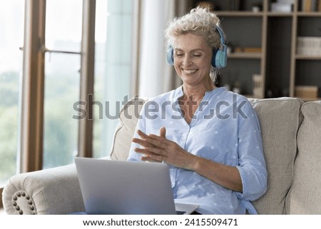 Happy aged woman in headphones sit on sofa with laptop watching on-line movie use subscription video streaming services, having pleasant videocall talk to family spend time on internet alone at home Royalty-Free Stock Photo #2415509471