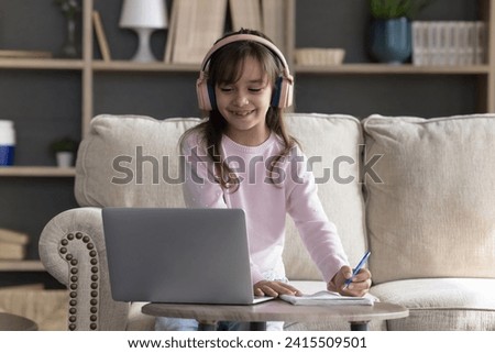 Cute preschooler girl in wireless headphones holding pen, take notes in copybook, e-learning, sit on sofa at table with laptop at home, listen tutor, enjoy remote education use videocall with teacher Royalty-Free Stock Photo #2415509501