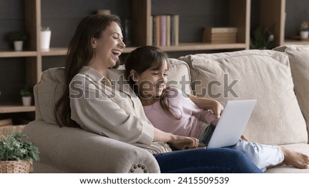 Woman and 6s daughter spend weekend watching comedy movies using laptop and digital online streaming services subscription, enjoy funny videos on internet relaxing together on cozy sofa in living room