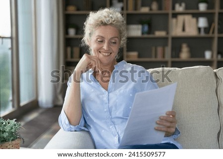 Smiling middle-aged woman read pleasant news in letter, sit on sofa alone at home, get paper notification from bank, get notice about loan repayment, pension rise, benefit. Achievement, finance growth Royalty-Free Stock Photo #2415509577