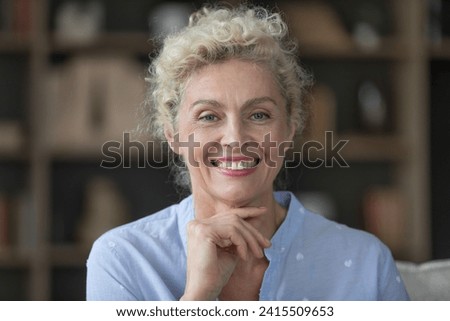 Close up head shot of beautiful mature woman smile, looking at camera relax alone on cozy couch in living room. Good-looking baby boomer generation female portrait, carefree retirement, natural beauty