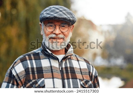 Handsome grandfather mature gray-haired man in a checkered shirt and glasses standing in a park in autumn. Royalty-Free Stock Photo #2415517303