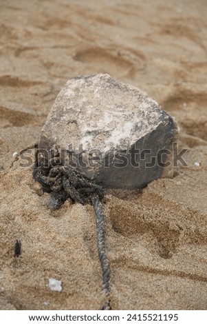 Exterior top view photo of an block of piece of rock stone on the sand of a beach used as an anchor as it's attached to a old damaged rope near the sea or ocean for a boat