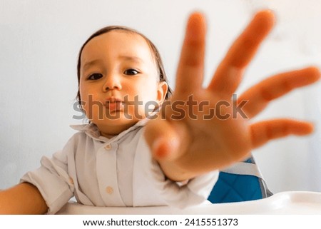 Face of Hispanic Latino baby sitting in white shirt raising left hand